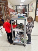 Three students in a school hallway gather around a rolling cart filled with books to choose their free prizes for Family Reading Week.
