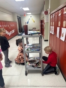 Three students gather around a rolling cart stacked with colorful books in a school hallway, selecting their free books for Family Reading Week.