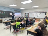 In a bright classroom, a female teacher stands at the front holding up an open picture book for her students. A group of elementary children sit at their desks, looking toward her with interest. The room is filled with instructional posters, a large digit