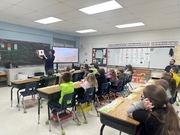 In a bright classroom, a female teacher stands at the front holding up an open picture book for her students. A group of elementary children sit at their desks, looking toward her with interest. The room is filled with instructional posters, a large digit