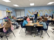 A teacher stands before a group of students seated at clustered tables in a blue-walled classroom. She holds a book open, showing the illustrations to the class. The wall behind her is decorated with a "Word of the Week" board and a creative student proje