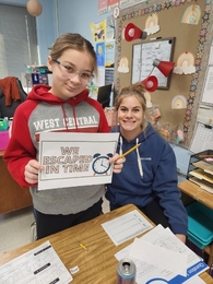 A girl in a red and grey "West Central" hoodie and a woman in a blue "Stetson" hoodie sit at a classroom desk covered in worksheets. The girl holds up a sign with an alarm clock icon that reads "WE ESCAPED IN TIME."
