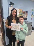 A woman in a black quilted vest and a boy in a light mint green hoodie stand by a classroom door. They are holding a sign that reads "WE CAME. WE SAW. WE ESCAPED."