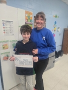 A woman in a bright blue sweatshirt stands behind a boy in a dark "Spyder" hoodie. They are smiling and holding a sign that reads "WE CRACKED THE CODE" in a classroom.