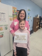 A woman in a pink sweater with white hearts stands behind a girl in a light pink hoodie. Both are smiling and holding a sign that reads "WE CAME. WE SAW. WE ESCAPED." in a bright classroom.