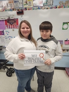 A woman in a white quarter-zip sweater and a boy in a "Cabela's" hoodie stand together in a classroom. They are holding a sign with a magnifying glass icon that reads "WE SOLVED THE CLUES."