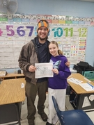 A man in an orange and grey winter hat and brown jacket stands smiling with a girl in a purple butterfly hoodie in a classroom. They are holding a white sign that reads "WE CRACKED THE CODE" with an illustration of a ring of keys.