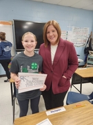 A woman in a maroon blazer stands next to a smiling girl wearing a grey 4-H t-shirt. The girl is holding a sign that reads "WE CRACKED THE CODE" in front of a classroom desk.