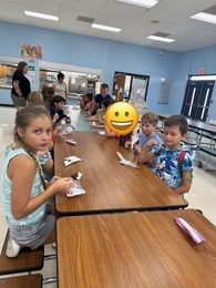 Elementary students sitting along long cafeteria tables during the summer reading party, smiling and eating ice cream bars and ice cream sandwiches together.
